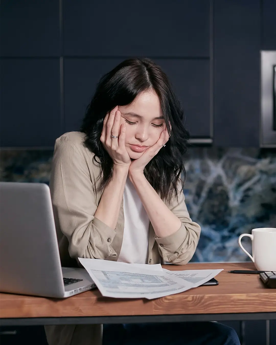 Picture of person overwhelmed at their desk with a laptop, a cup of coffee, and reports in front of them.