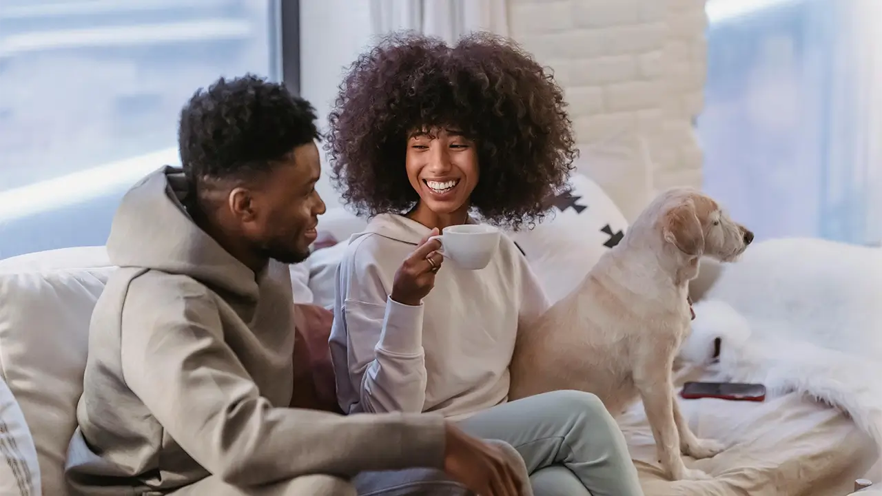 Man, woman and their dog sitting on their couch. The woman is enjoying a cup of coffee and laughing.