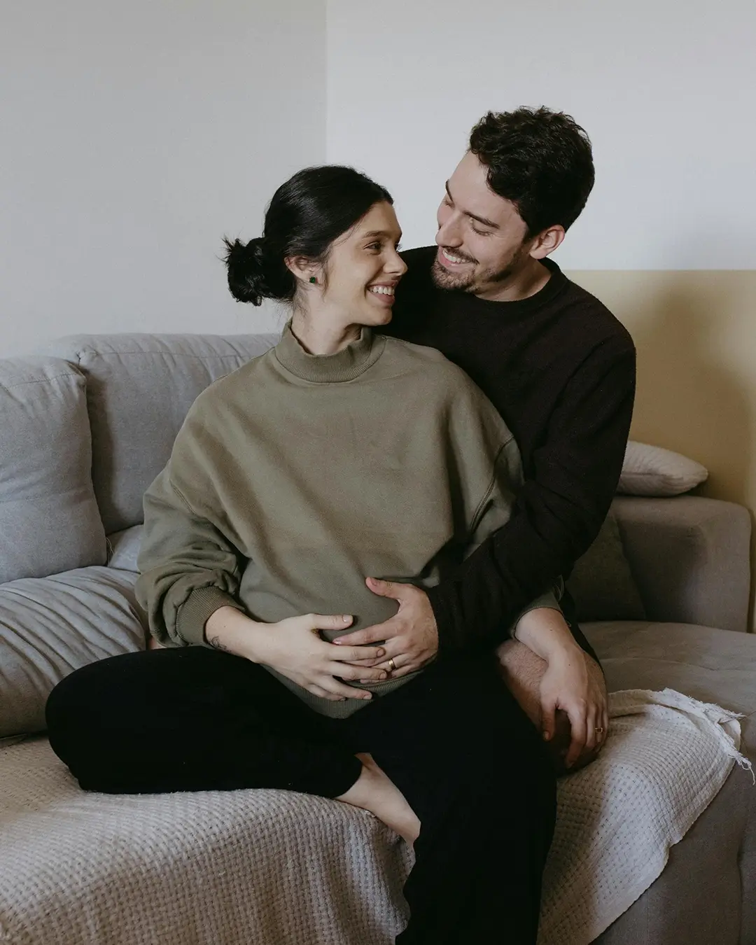 Man and pregnant woman cuddling on their couch smiling at each other.